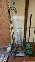 Photo showing tall black metal plant hangers, wire garden cage, bird decorations, and garden tools on the floor against the wall.