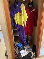 Hein Gericke yellow and purple rain suit and Aerostich RoadCrafter red protective suit jacket and pants hanging side by side; various motorcycle gear on floor below includes gloves and bags.