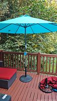 Full view of the blue outdoor umbrella set up on a patio, accompanied by red cushioned outdoor furniture and the black metal umbrella base stand on the deck floor.