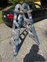 Multi-position aluminum ladder set up in an A-frame position on an outdoor gravel and driveway surface with a black car in the background.