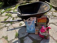 Full view of black metal wheelbarrow with the five bags arranged in front: Diamond Crystal de-icer, Ye Melt ice melt, Supersoil garden sand (two small bags), and bright pink Outdoor Potting Soil bag.