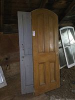 Two antique wooden doors leaning against a wall inside a shed or attic space. The door with the rounded top is natural wood color, positioned in front of the gray flat-top door. Both doors show signs of aged wear and patina.