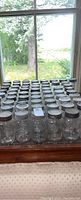 62 clear glass canning jars arranged in rows on a window ledge, showing glass and metal lids and embossed logos.