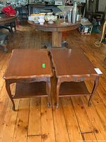 Pair of wooden end tables with faux drawers and lower shelves shown side by side on wood plank floor with visible wear and scuffs.