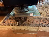 Clear glass side table placed on a patterned rug with a TV stand on top, showing the table's transparency and back power cords visible through it.