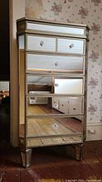 Front view of mirrored chest showing five drawers and acrylic knob pulls on a hardwood floor against floral wallpaper.