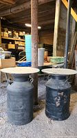 Pair of black vintage metal milk cans with round acrylic tabletops, placed side by side on gravel floor, showing significant corrosion and paint wear.