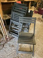Set of several folded dark wood patio chairs leaning against a surface with one unfolded chair in front on gravel floor.