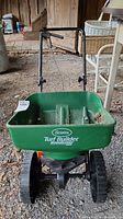 Front view of Scotts Turf Builder Edgeguard Mini seed spreader showing green plastic hopper with brand logo and large black wheels on gravel surface inside a garage area.