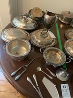 Silverplate serving dishes including covered oval dishes, pitcher, lids, and small utensils arranged on a wooden table showing tarnish and wear.
