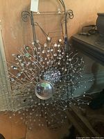 Metal sunburst mirror with curved designs tipped with small reflective spheres, leaning against a wall next to a plate holder.