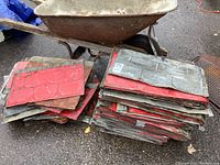Full view of stacks of old tin ceiling tiles, showing red paint wear and rust, placed outdoors on asphalt near a rusty wheelbarrow