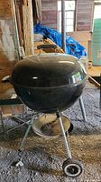 Front view of black round Weber charcoal grill kettle on metal legs with wheels, showing the grill body and attached metal base.