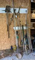 Full view of various yard tools leaning against a wooden wall on a gravel floor showing pitch forks, shovels, hoes, rakes, and clippers.