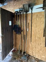 Photo of yard tools hanging on plywood wall including shovels, rakes, pick axe, stamper, and hoe all showing signs of heavy use and rustic condition.