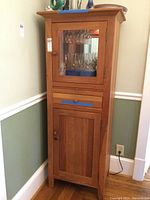 Full view of tall wooden bar cabinet with glass door and drawer. Shows cabinet's exterior features and wood finish.