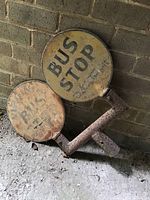 Two circular vintage bus stop metal signs leaning against a wall. Both show significant rust and fading.
