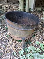 Side view of large metal cauldron outdoors, showing three legs and wide rim with rust.