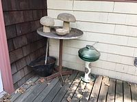 Wide view of corner showing all items: three concrete mushrooms on wooden table, black coal bucket beneath, green and white bird feeder to right, placed on wooden deck against house corner with siding.