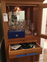 Wooden cabinet with upper display area holding assorted crystal stemware including glasses, decanter, and a closed drawer below with bar accessories inside