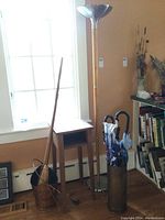 Overview of the floor lamp, wooden end table, umbrella stand with umbrellas, brooms, and metal bucket in a corner of a room.