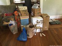 Photo showing stacks of boxes, paper bags with pinecones and steel wire, a scarecrow decoration, and a brass reindeer candle holder with original foam packaging.