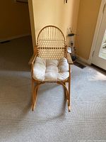 Front-facing view of natural wood rocking chair with woven backrest and off-white seat cushion on carpeted floor.