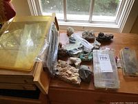 Wide view showing multiple rock and gem specimens spread on a wooden desk including large raw rocks and multiple plastic storage cases.