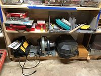 View of shelving with multiple small cases and boxes above, power tools (compound miter saw, circular saw, sander) below, located in basement space.