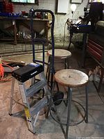 Wide view of two metal bar stools with round cream seats, Husky two-step aluminum stepladder, and blue hand truck in basement setting.