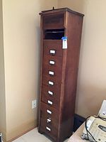 Front view of tall narrow oak file cabinet with eight drawers against a beige wall