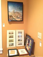 View of wall with large desert landscape painting and several framed pieces leaning against the wall.