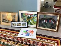 Five framed artworks arranged leaning against a wall on a carpeted floor, showing a range of styles and sizes.