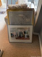 Two framed prints leaning against wall: front color tools scene, behind gold-framed figurative print