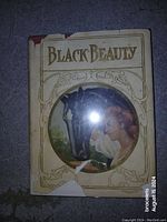Front cover of Black Beauty vintage book from 1908 with decorated dust jacket featuring a woman and horse illustration, titled 'Black Beauty'. Shows signs of wear with some tearing and discoloration.