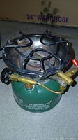 Green metal camp stove with black burner grate and brass controls on a concrete floor.
