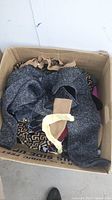 View into an open cardboard box filled with folded and crumpled women's clothing including a dark gray sweater and a patterned garment.