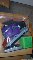Photo of items sealed in a plastic bag including a purple handheld device, wired headset microphone, pink game console, a small grey radio speaker, and a green digital clock.