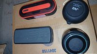 Four portable speakers arranged on a cardboard surface, including JBL, JVC, and two unidentified rectangular models.
