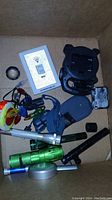 View of box containing various flashlights, a gray electronic light switch panel, black plastic mount, and miscellaneous components.