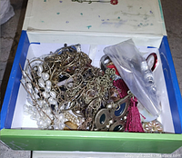 Top view of an open box containing various pieces of mixed jewelry including chains, faux pearl accessories, tassel earrings, and brooches.
