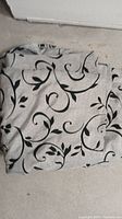 Light-colored curtain with black leaf and swirl pattern folded on floor.