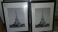 Two black and white Eiffel Tower pictures side by side in black frames with white mats, leaning against a wall.