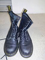 Pair of black Dr. Martens boots standing open on floor, showing leather upper, eyelets, laces, and branded heel pull tab