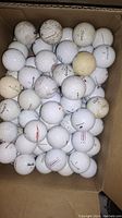Top view of a cardboard box filled with over 50 used golf balls, some with visible brand markings and discoloration.