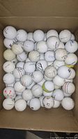 Photo of a box filled with approximately 50 mixed golf balls, predominantly white, showing various brands and signs of wear.