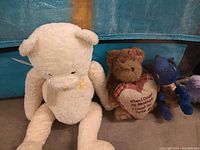 Photo showing a large cream colored stuffed bear with a bow tie, a brown teddy bear holding a heart pillow with text, and a small blue plush cat.