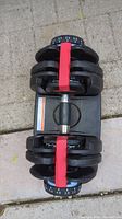 Top view of adjustable dumbbells mounted in black base with visible red handle and weight plates.