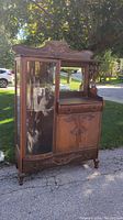 Front view of the antique buffet cabinet showing the wooden top with carved details, curved glass display section on the left, drawer, and cabinet doors on the right.