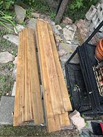 Multiple cedar wood planks stacked on the ground near garden and outdoor shelving. Shows length and width of planks.